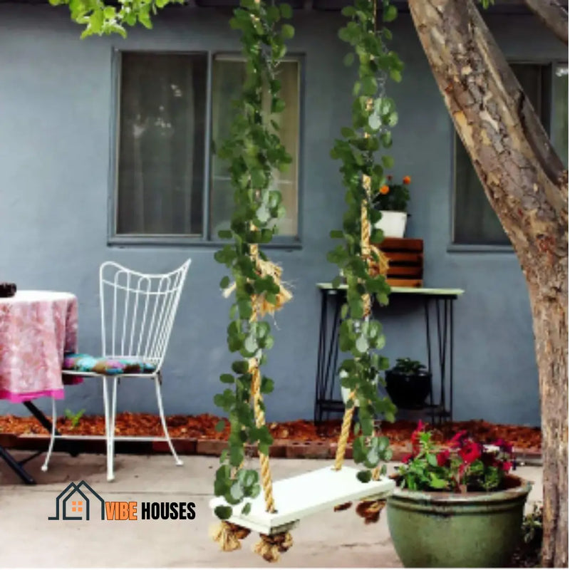 Artificial plants - Image of a swing attached to a tree. The ropes of the swing are intertwined with begonia leaves. In the background a blue house. Vibe Houses 